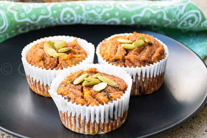 Three paleo pumpkin muffins in white paper liners, topped with pepitas, on a black plate.