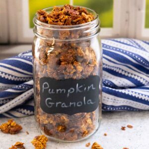 Glass jar labeled Pumpkin Granola filled with chunky granola, set on a gray surface near a window with a striped cloth in the background.