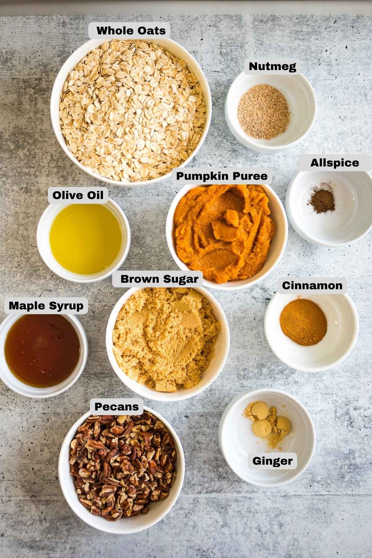 Overhead view of labeled ingredients in white bowls, including whole oats, pecans, pumpkin puree, brown sugar, maple syrup, olive oil, nutmeg, allspice, cinnamon, and ground ginger.