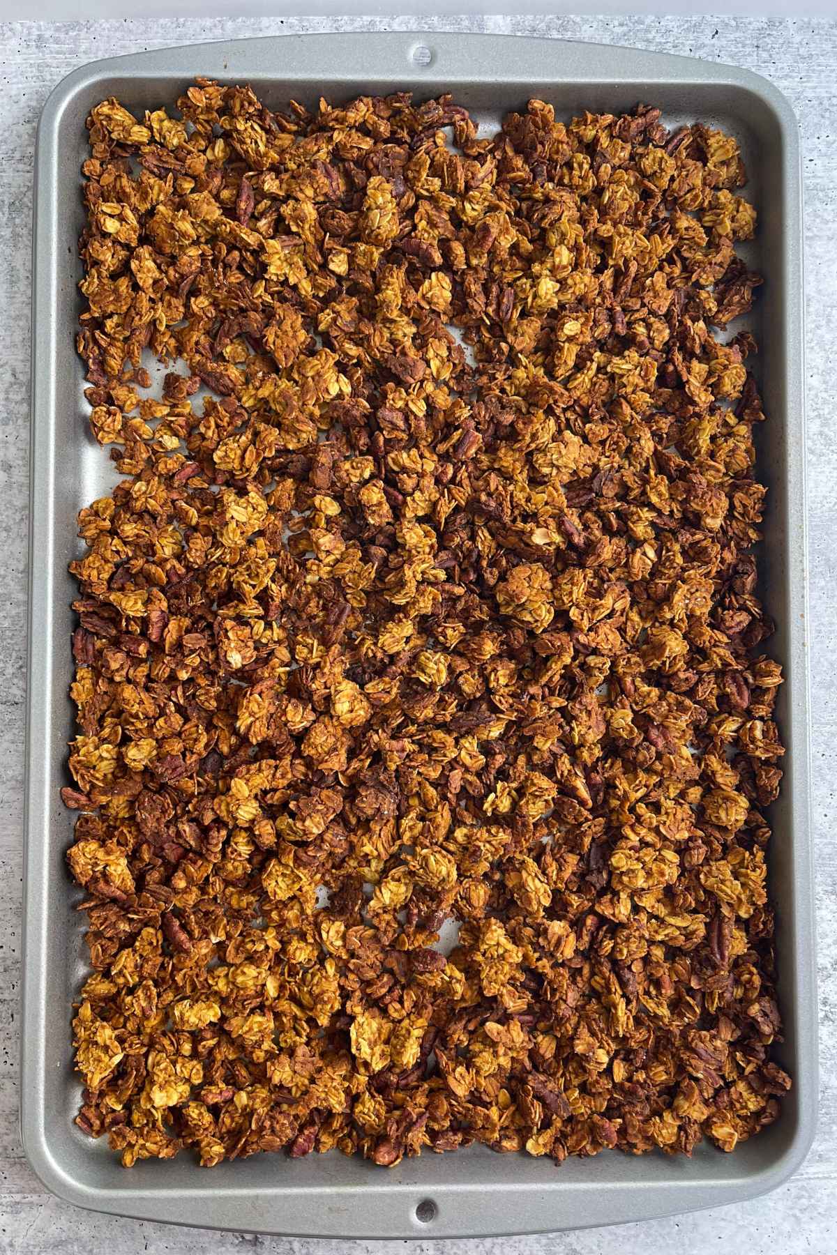 Overhead view of freshly baked pumpkin granola spread out on a metal baking sheet, with golden brown clusters visible.