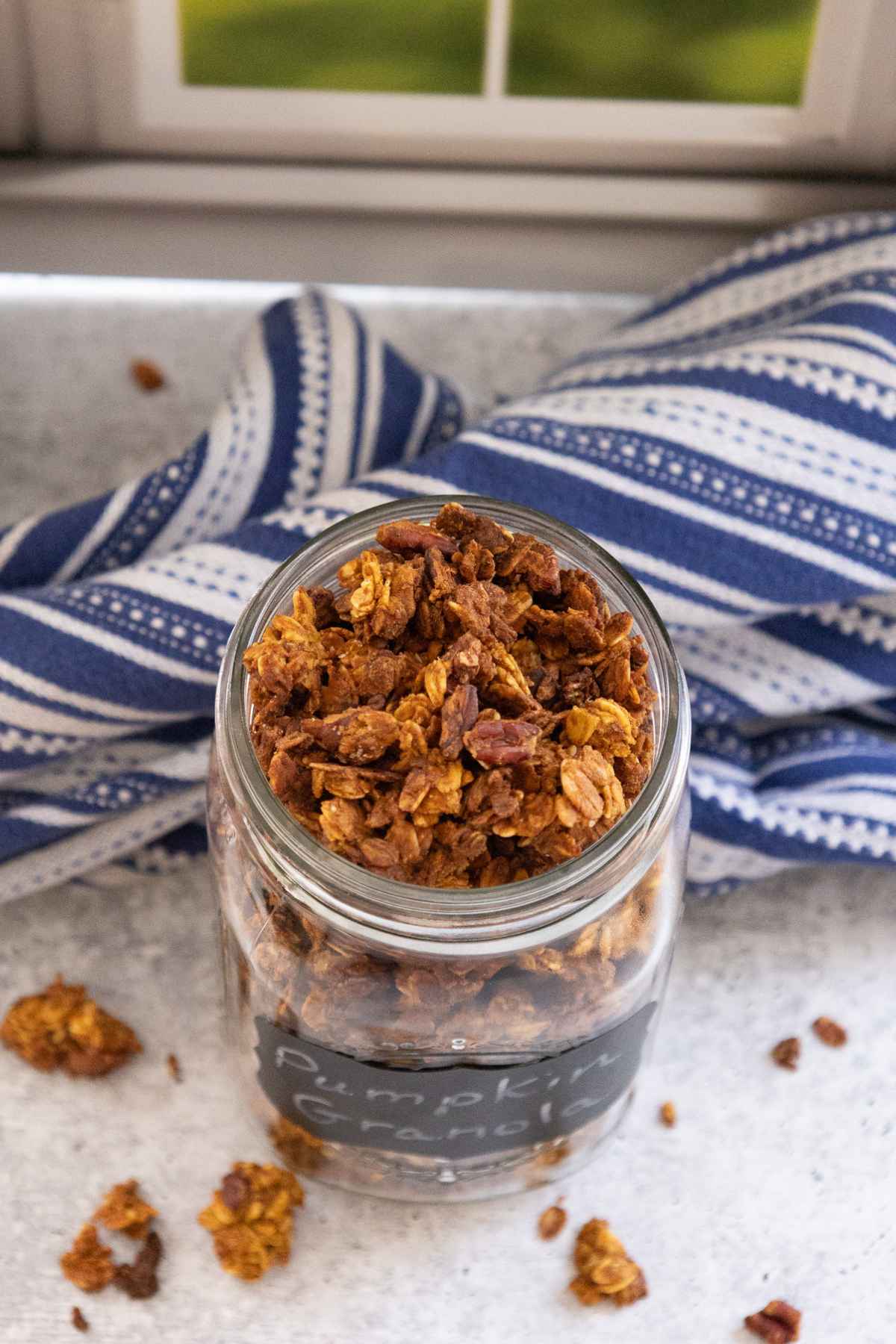 Overhead view of freshly baked pumpkin granola in a glass jar placed in front of a blue and white striped cloth with golden brown clusters scattered around the jar.