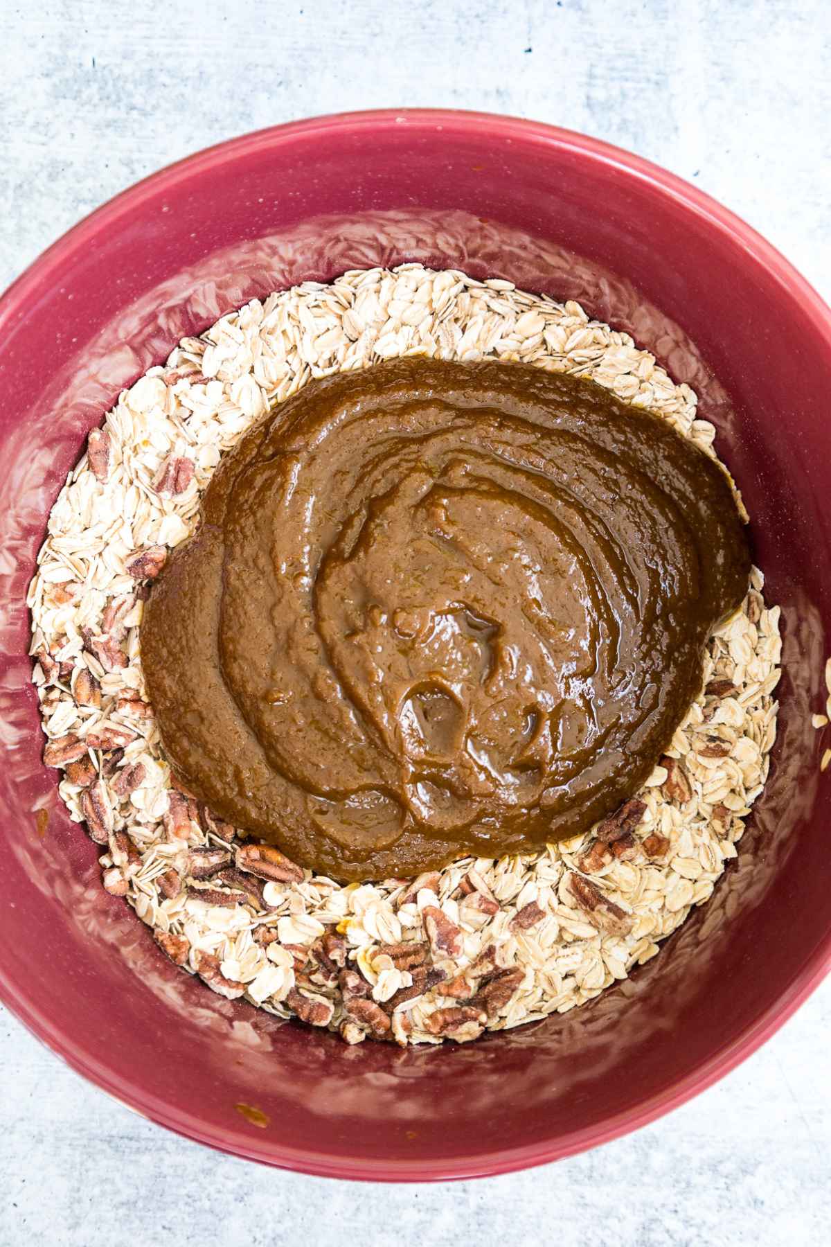 Large red mixing bowl filled with whole oats and chopped pecans, topped with a thick brown pumpkin mixture, ready to be stirred together.