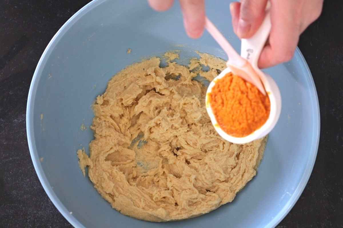 Measuring cup filled with pumpkin puree being added to a bowl of cookie dough.