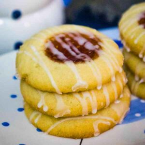 A stack of three raspberry thumbprint cookies on a plate, each topped with jam and a drizzle of icing.