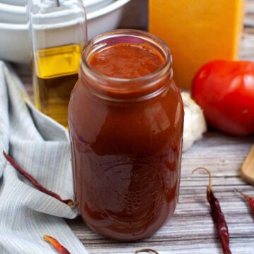 A glass gar of red enchilada sauce on a wooden surface with olive oil, cheese, tomato, and garlic blurred in the background.