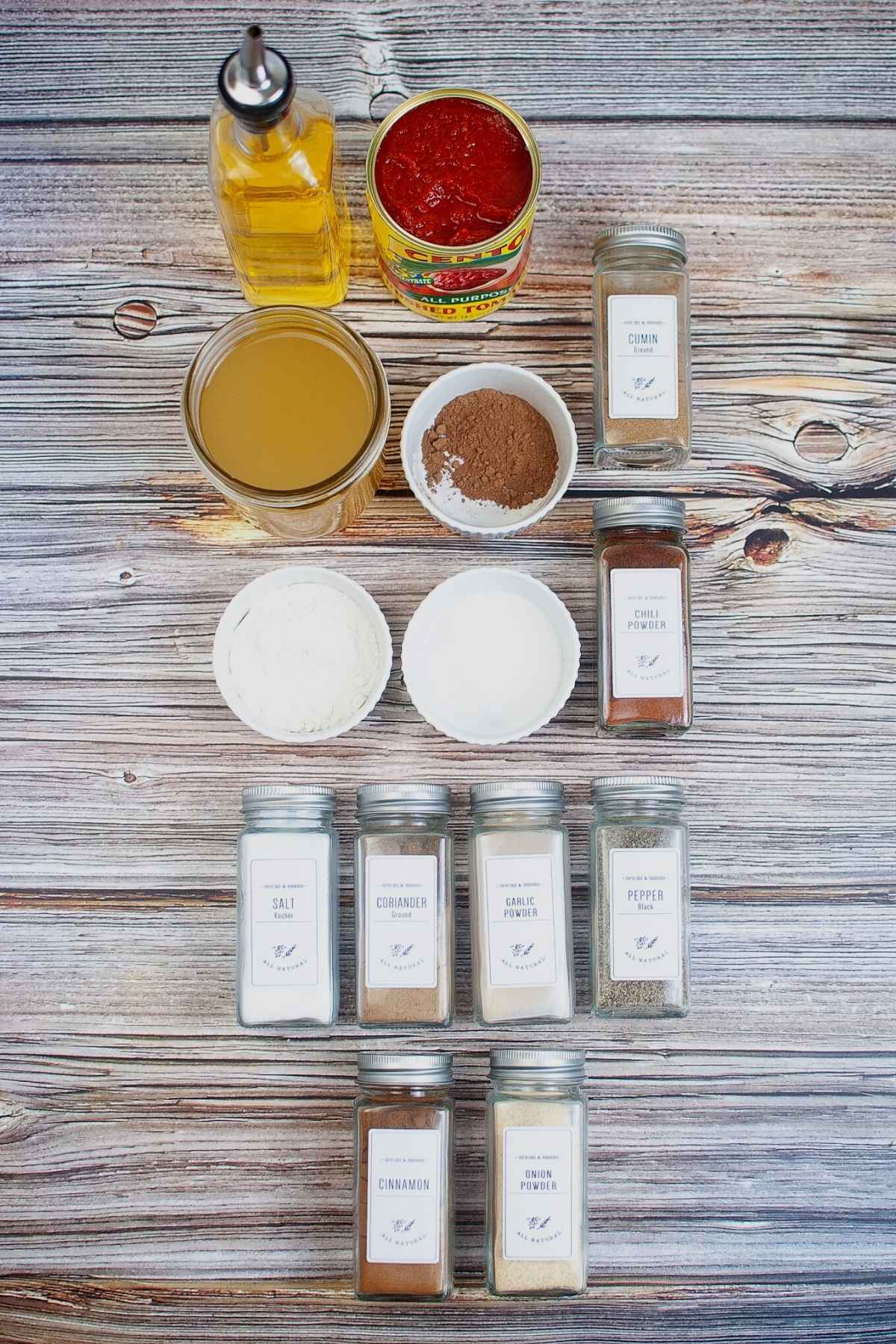 Overhead layout of red enchilada sauce ingredients including canned tomatoes, broth, oil, flour, cocoa powder, and an assortment of labeled spices in jars.