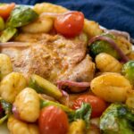 A white plate with roasted chicken thighs served with golden gnocchi, brussels sprouts, cherry tomatoes, and red onions on a plate.