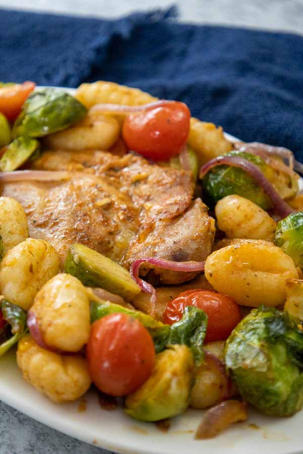 A white plate with roasted chicken thighs served with golden gnocchi, brussels sprouts, cherry tomatoes, and red onions on a plate.