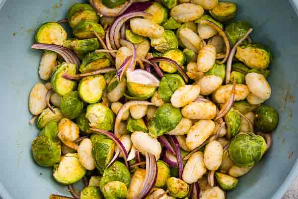 A mixing bowl filled with Brussels sprouts, gnocchi, and red onion slices tossed in seasoning.