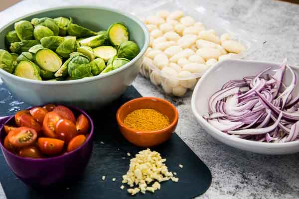 A display of ingredients including Brussels sprouts, gnocchi, red onion slices, cherry tomatoes, minced garlic, seasoning, and olive oil.