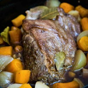 Slow cooker filled with cooked cchuck roast, tender carrots onions, and potatoes simmering in broth with bay leaves.