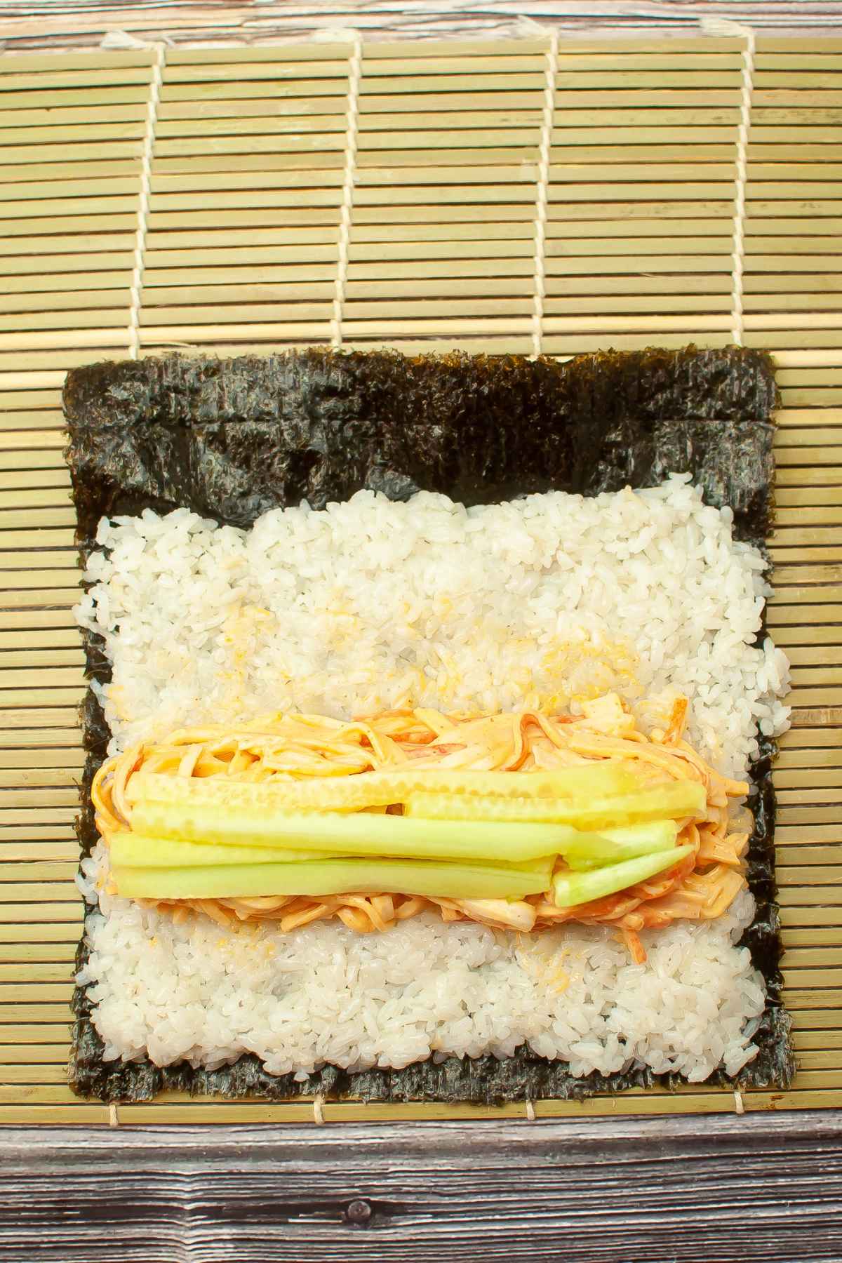 A sheet of nori on a bamboo mat topped with sushi rice, shredded crab mixture, and cucumber sticks ready to roll.