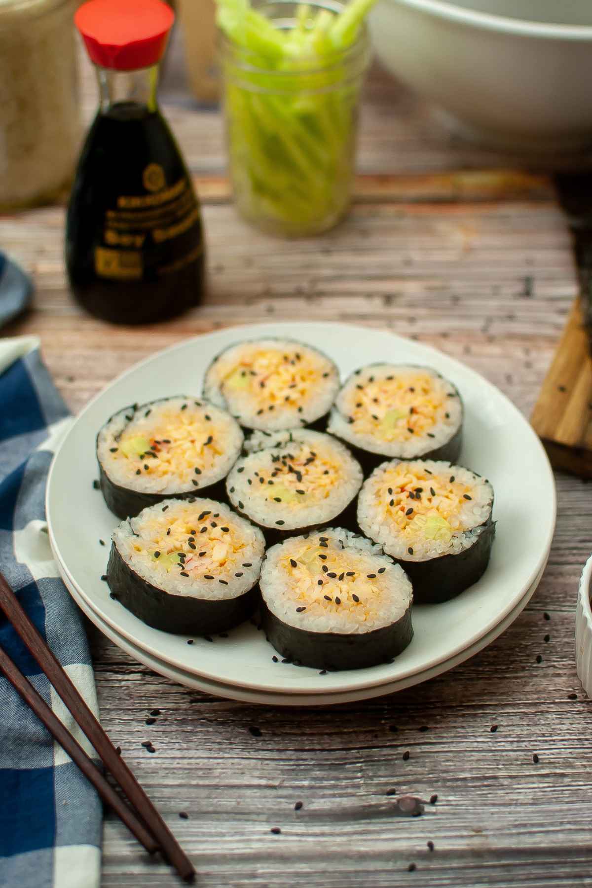 A white plate with sliced spicy crab sushi rolls filled with crabmeat and cucumber, sprinkled with black sesame seeds, served with soy sauce in the background.