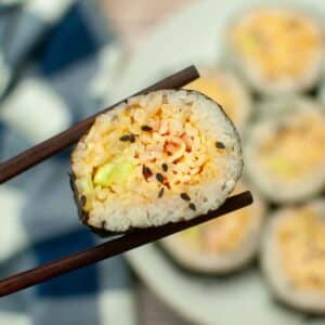 A piece of spicy crab sushi roll held with chopsticks, showing cucumber, rice, and crab filling with sesame seeds.