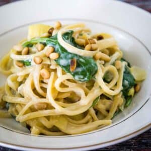 Image of creamy spinach artichoke pasta in a cream gold rimmed bowl garnished with toasted pine nuts.