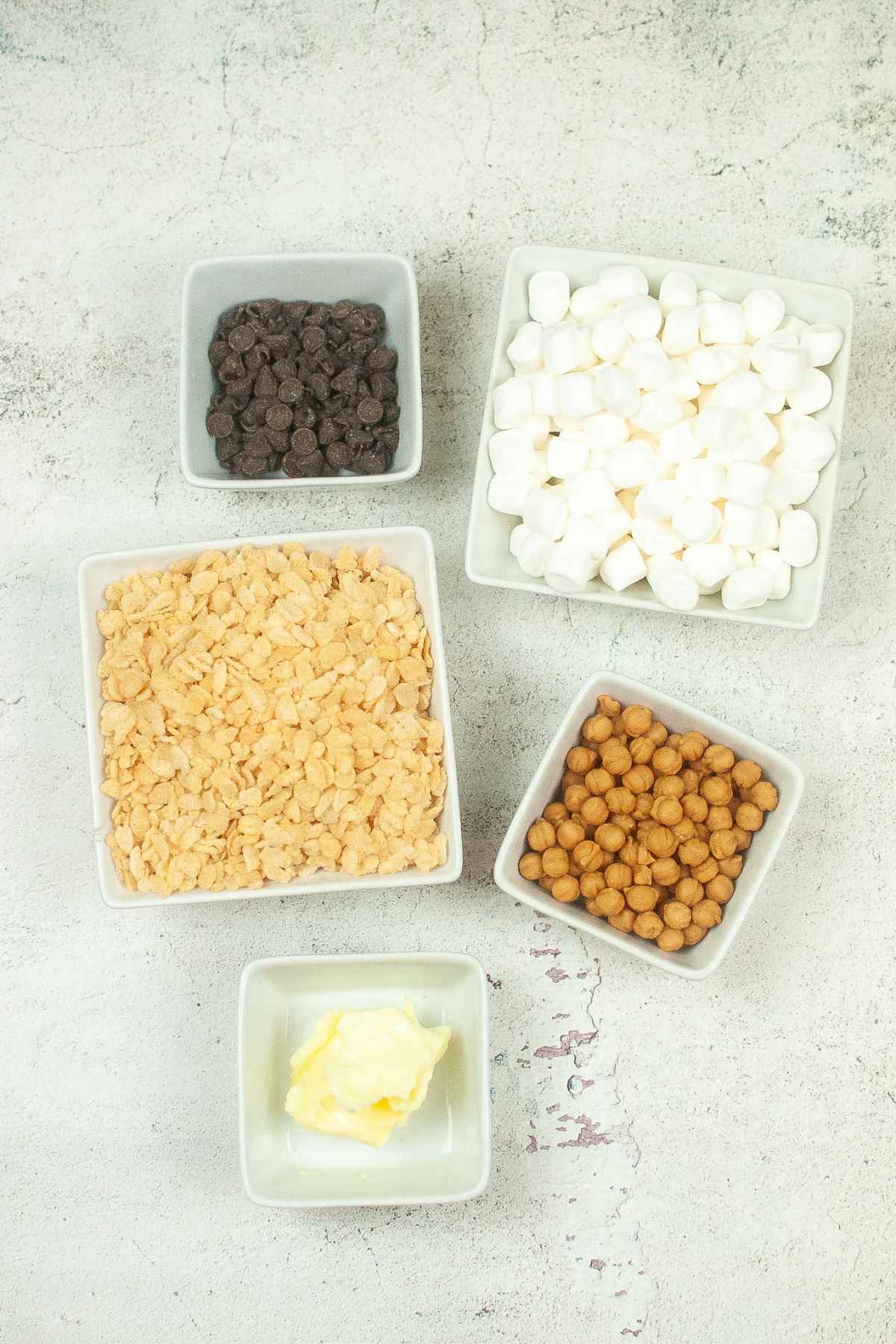 Overhead view of ingredients in white bowls, including chocolate chips, marshmallows, rice crispies, caramel bits, and butter.