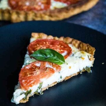 Slice of tomato pie with cheese filling and fresh basil and tomato slices on a black plate.