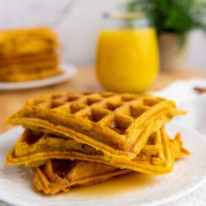 Close up of pumpkin waffles stacked on a white plate, drizzled with syrup, next to a glass of orange juice on a white textured cloth.