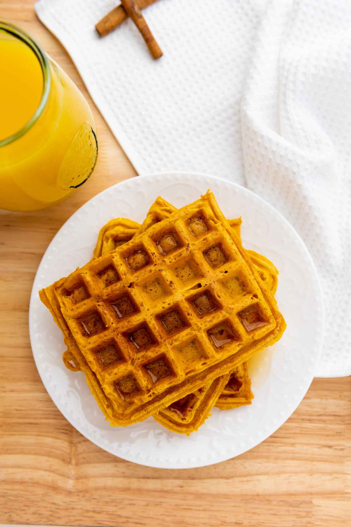 Overhead view of a stack of golden pumpkin waffles on a white plate, served with a glass of orange juice and a white textured napkin on a wooden surface. Two cinnamon sticks rest in the background.