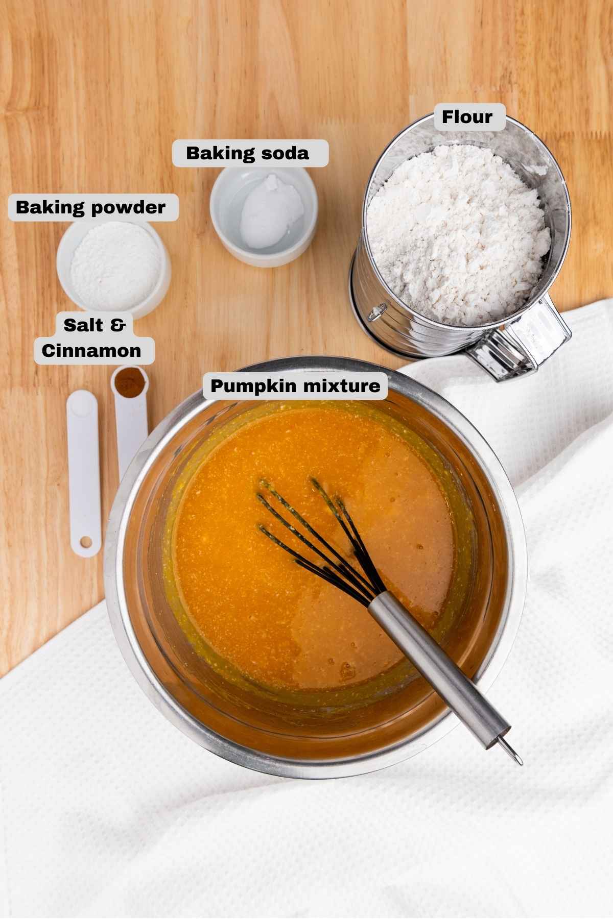 Flat lay of ingredients on a wooden surface, including a bowl of pumpkin mixture with a whisk, a sifter filled with flour, and small bowls, containing baking powder, baking soda, salt, and cinnamon.