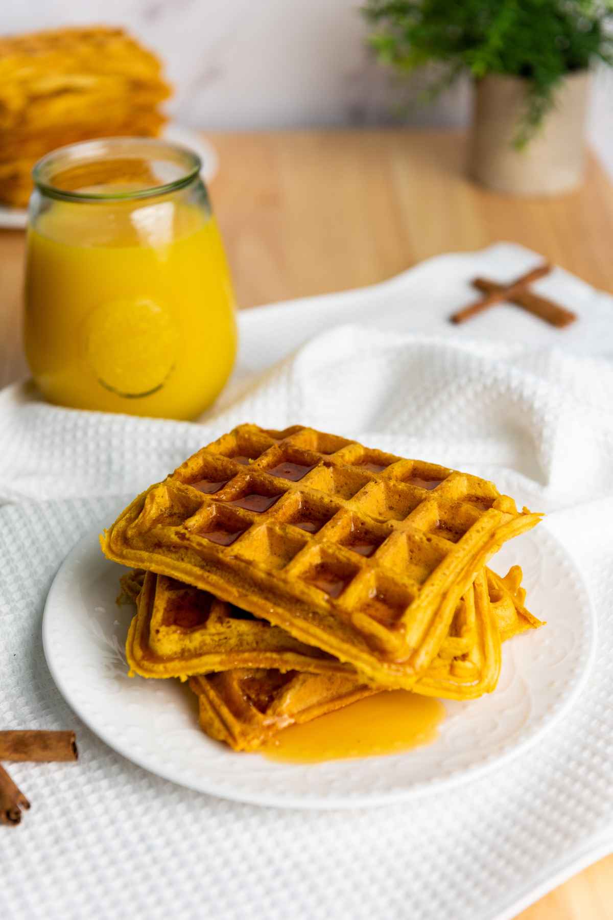 Close up of pumpkin waffles stacked on a white plate, drizzled with syrup, next to a glass of orange juice on a white textured cloth.