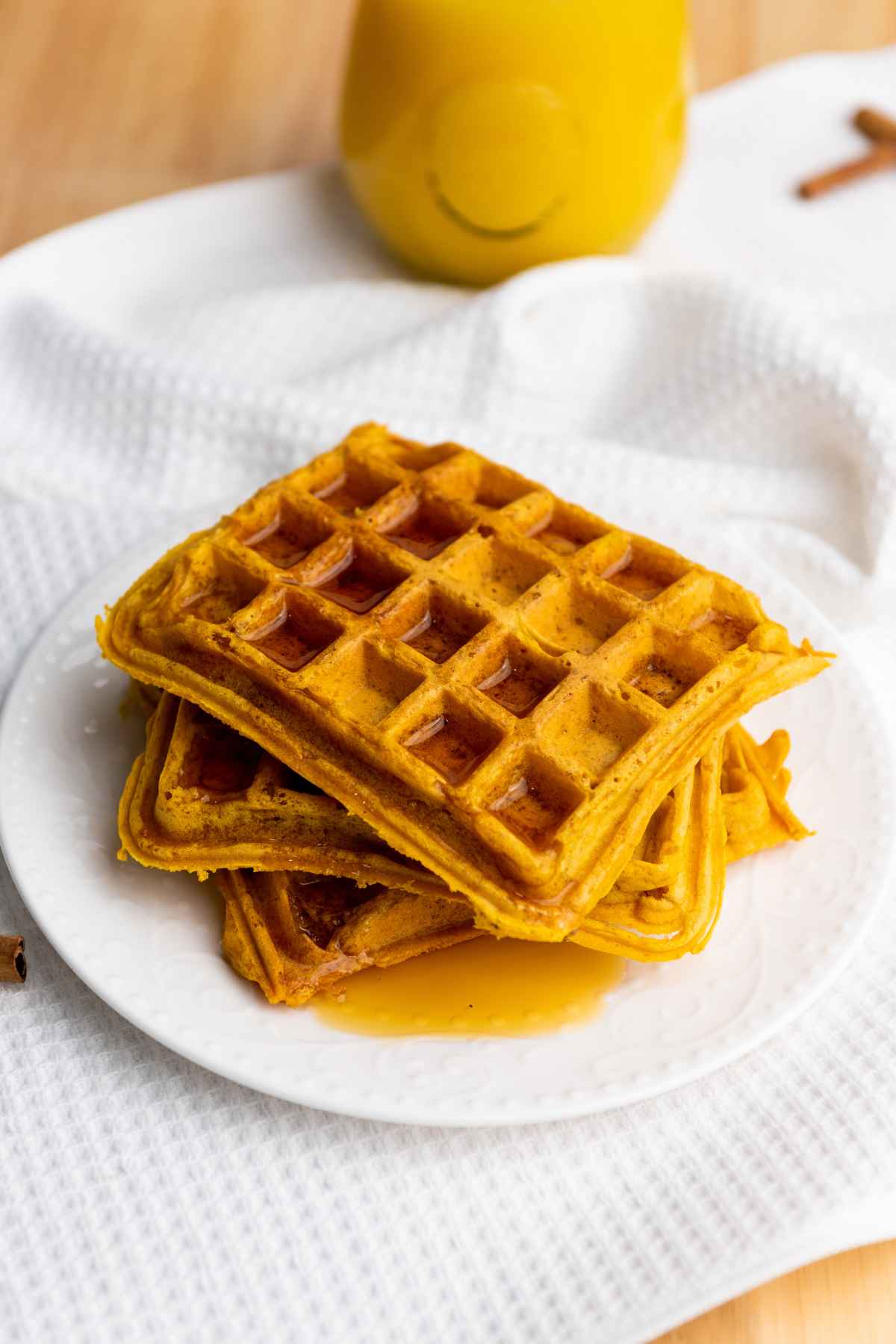 Close up of pumpkin waffles stacked on a white plate, drizzled with syrup, next to a glass of orange juice on a white textured cloth.