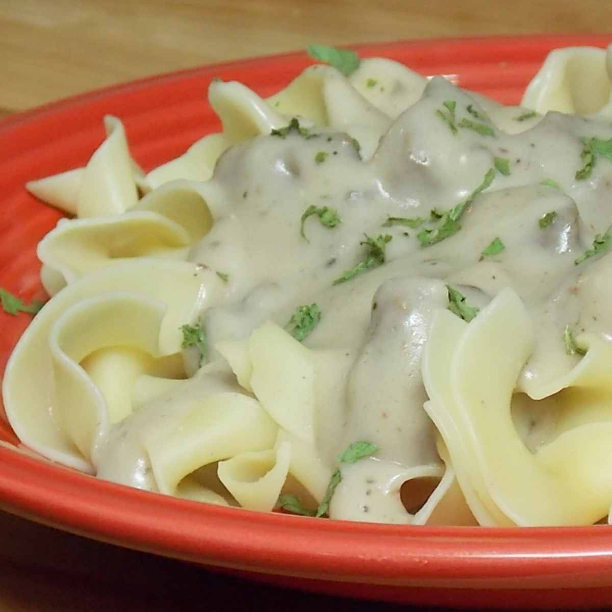 Wide egg noodles topped with a venison and mushroom gravy sprinkled with parsley served on a red plate.