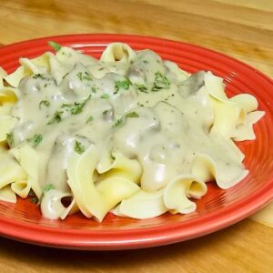 Wide egg noodles topped with a venison and mushroom gravy sprinkled with parsley served on a red plate.