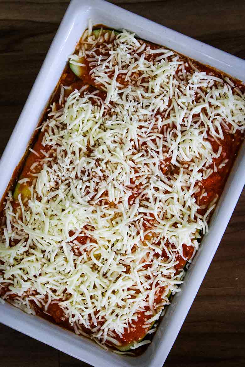 A white baking dish filled with zucchini noodle lasagna layered with sauce and generously covered in shredded mozzarella cheese, ready for baking.