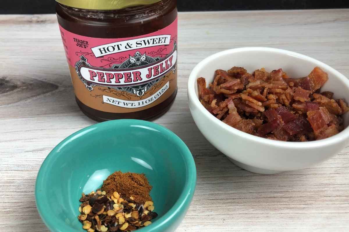 A jar of hot and sweet pepper jelly, a white bowl filled with crispy bacon, and a turquoise bowl with red pepper flakes and cayenne pepper.
