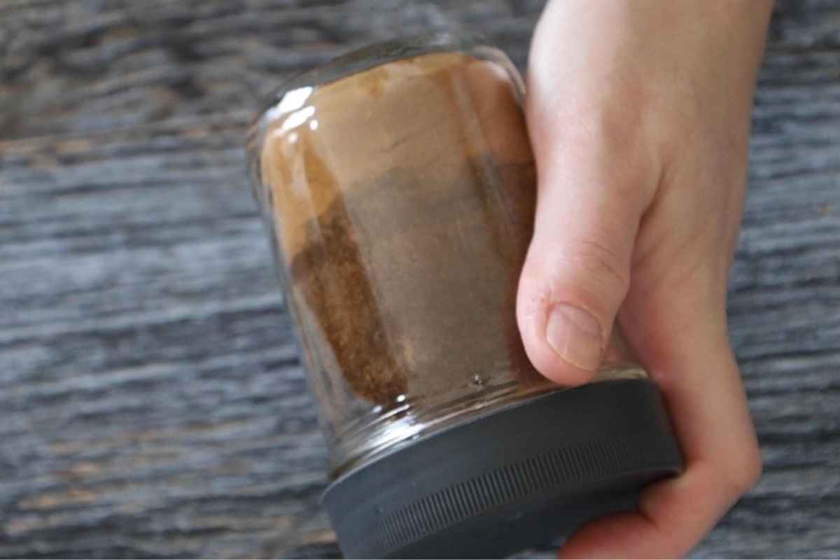 A hand holding a glass jar of brown spices showing through the glass.