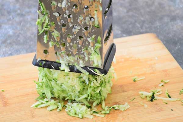 Freshly grated zucchini collected under a metal box grater on a wooden cutting board.
