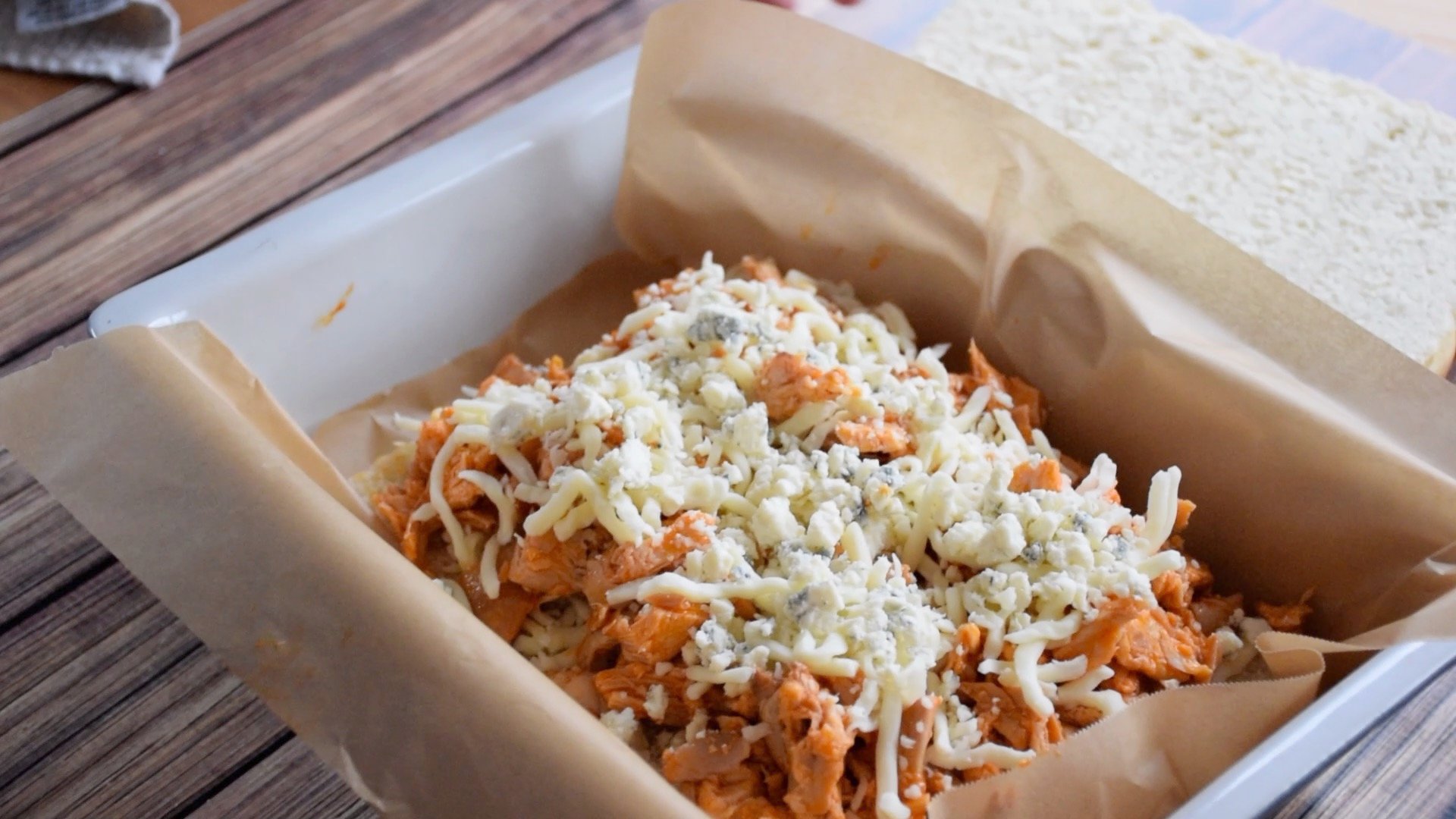 Shredded buffalo chicken and a mix of shredded cheese layered on the bottom half of slider buns inside a parchment lined baking dish.