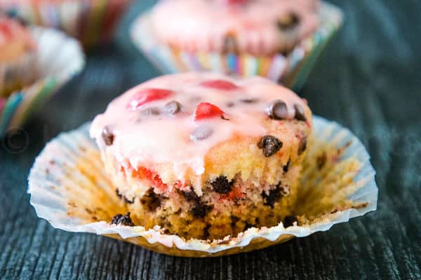 A chocolate chip muffin in a partially wrapped striped liner, showing the pink glaze, cherries, and chocolate chips.