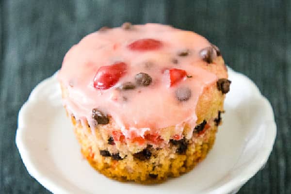 A close up of a cherry chocolate chip muffin topped with pink glaze, sitting on a white plate.