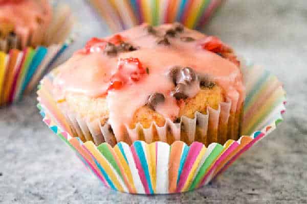 A cherry chocolate chip muffin in a colorful striped paper liner, topped with pink glaze and chocolate chips.