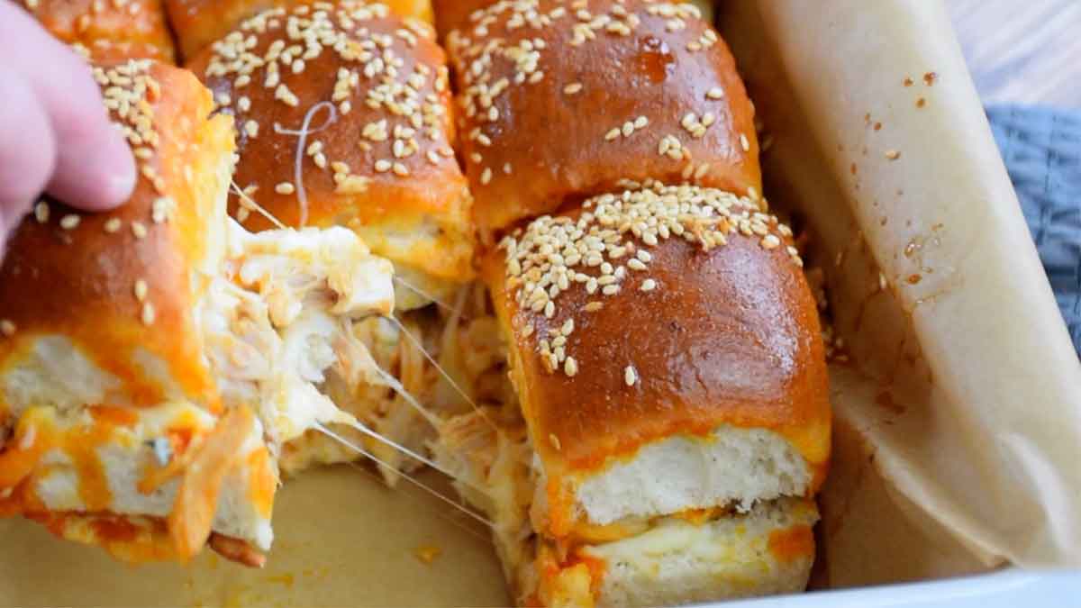 A hand lifting a cheesy buffalo chicken slider from a baking dish, with melted cheese stretching between the rolls.