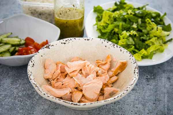 Ingredients for Greek salmon salad, including cooked flaked salmon, lettuce, cucumbers, tomatoes, and dressing.