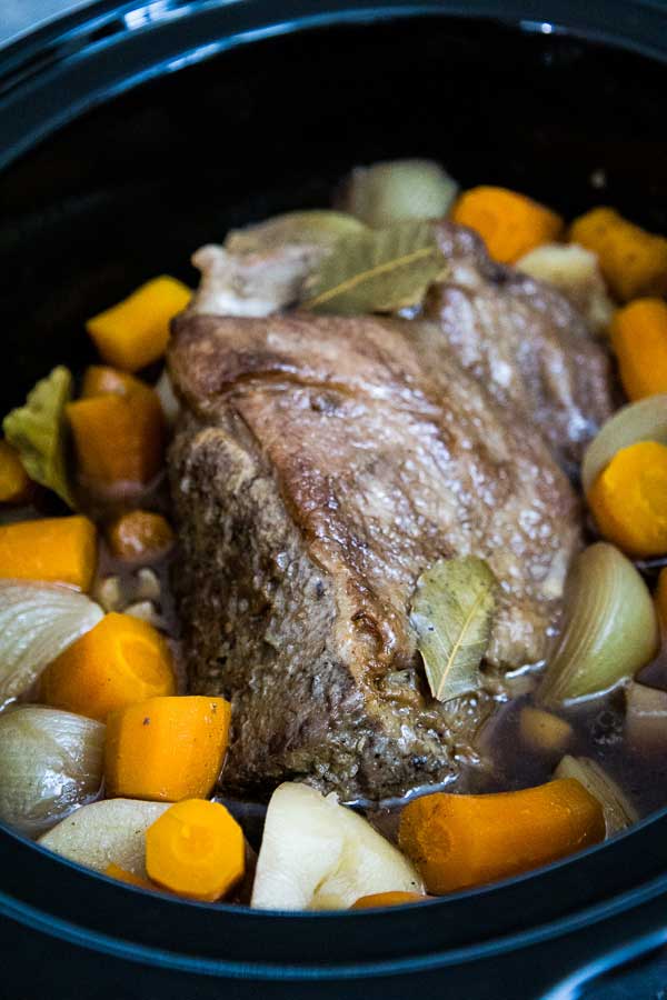 Slow cooker filled with cooked chuck roast, tender carrots onions, and potatoes simmering in broth with bay leaves.