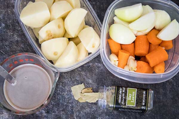 Ingredients for roast: peeled potatoes, chopped onions, carrot pieces, whole garlic cloves, a measuring cup of broth, and a jar of bay leaves.