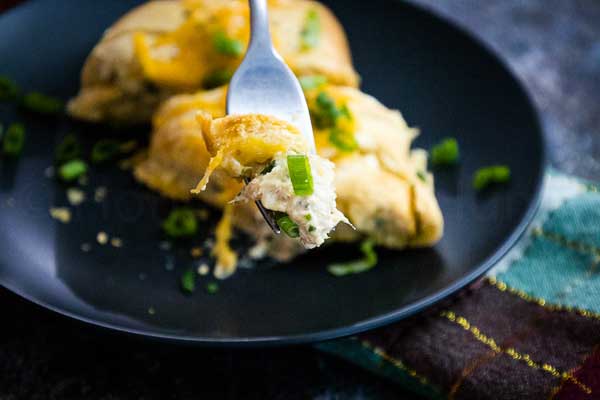 A fork lifts a bite of cheesy tuna crescent braid topped with melted cheddar and fresh green onions from a black plate.