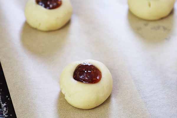 Unbaked thumbprint cookie dough with jam filling on a parchment lined baking sheet.