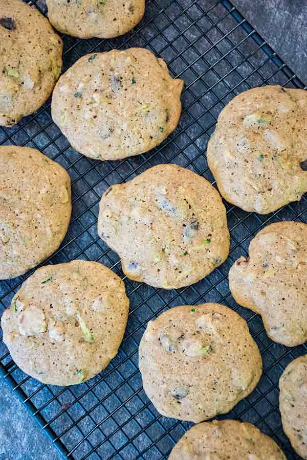 Zucchini spice cookies cooling on a wire rack, each dotted with visible pieces of zucchini and nuts.