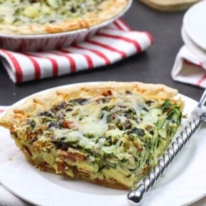 A single slice of bacon mushroom quiche on a white plate with a fork, with the rest of the quiche in the background.