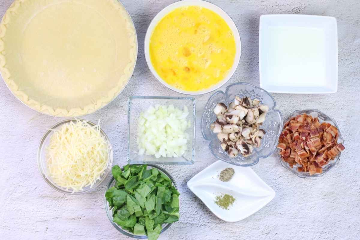 Overhead view of ingredients including eggs, milk, cheese, spinach, onion, mushrooms, bacon, seasonings, and an unbaked pie crust.