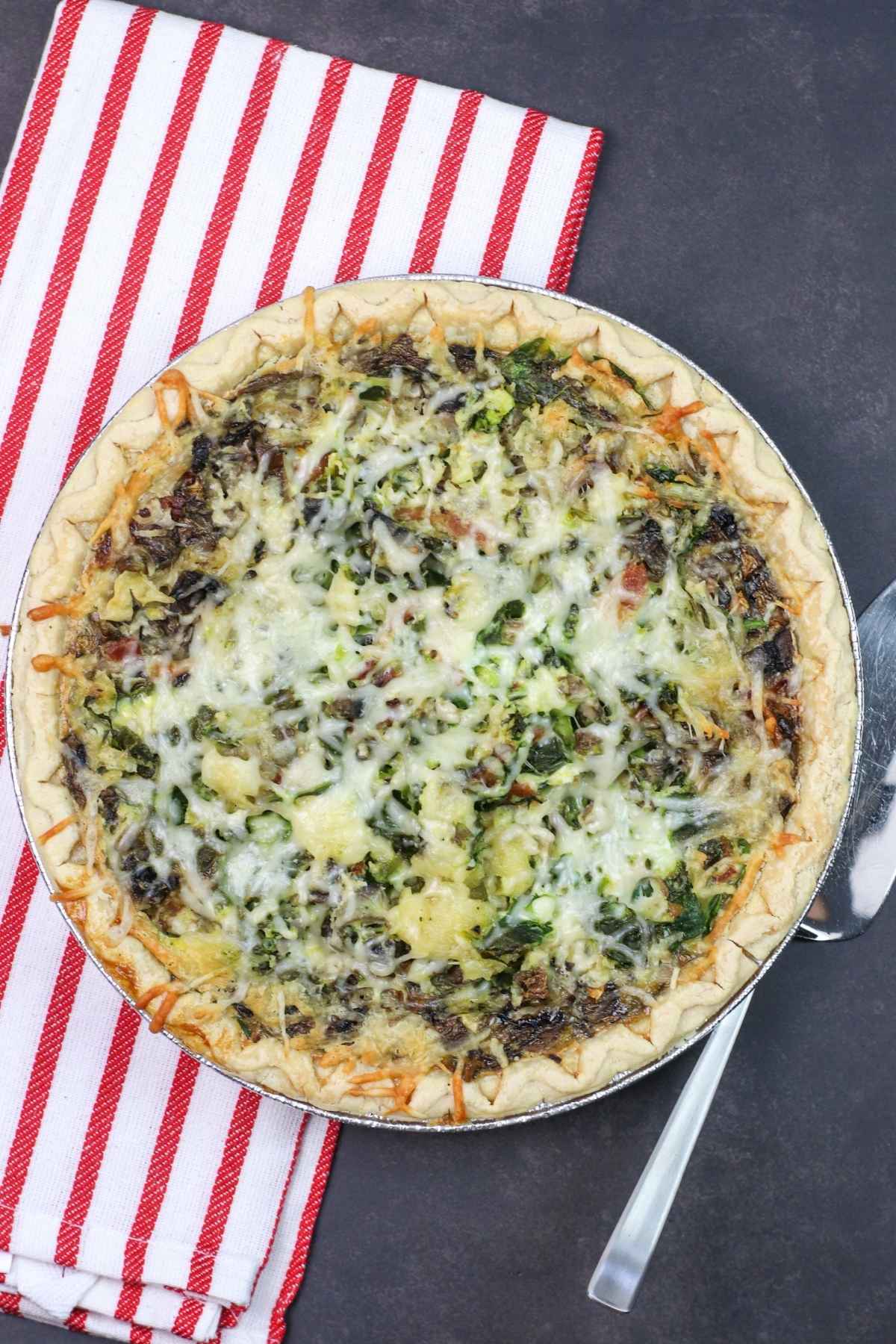 Overhead view of a whole baked bacon mushroom quiche in a pie pan, topped with melted cheese.