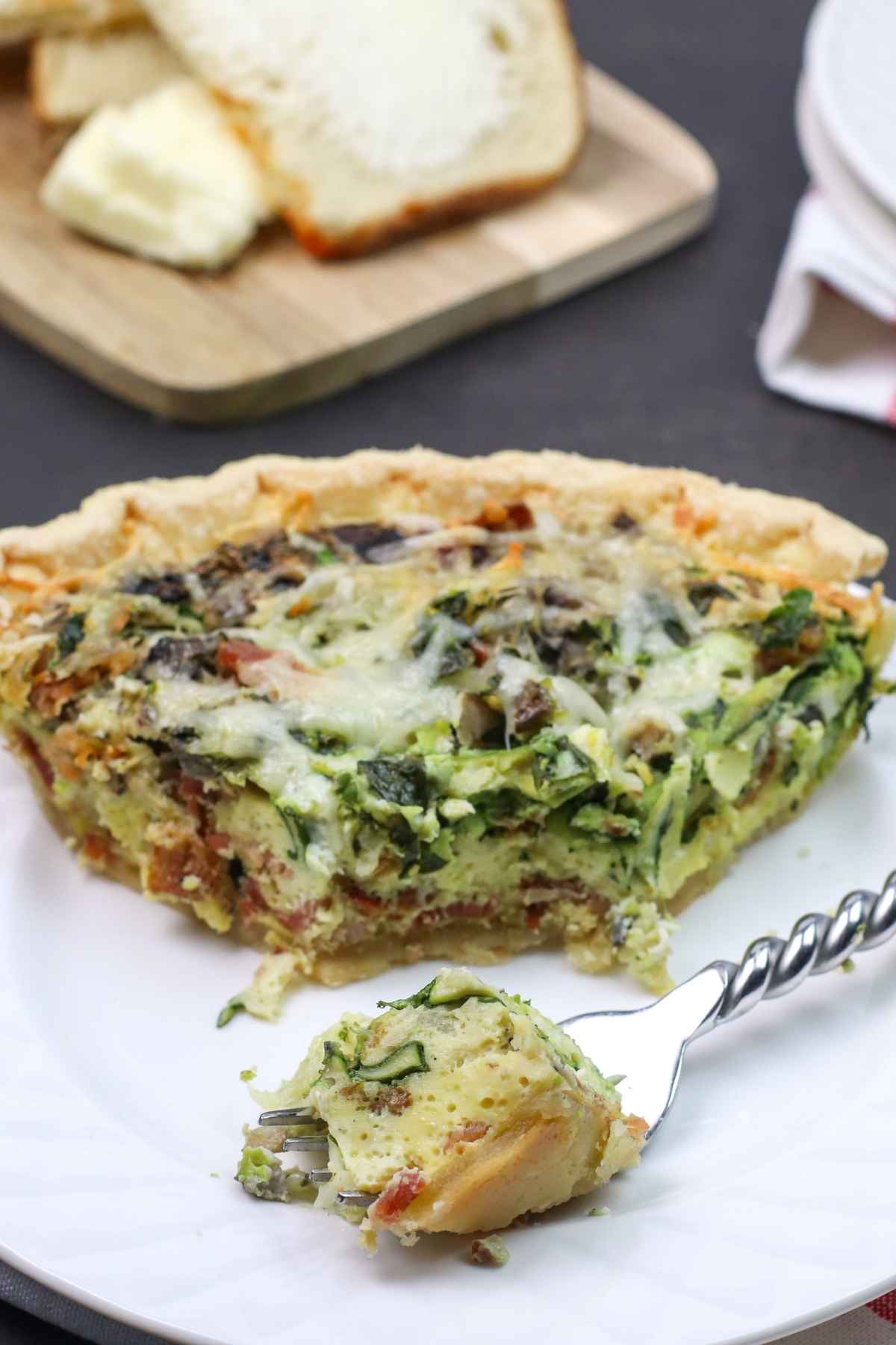 A slice of bacon mushroom quiche on a white plate, with a fork holding a bite sized piece in the foreground.