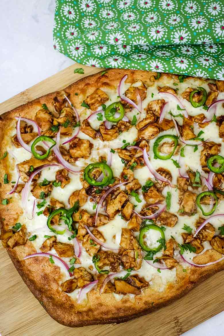 Overhead of keto bbq chicken pizza resting on a wooden board with a green and white floral napkin next to it.