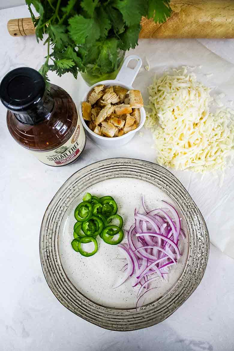 Overhead image of pizza toppings including sliced jalapenos, red onions, bbq sauce, chicken, and mozzarella cheese.