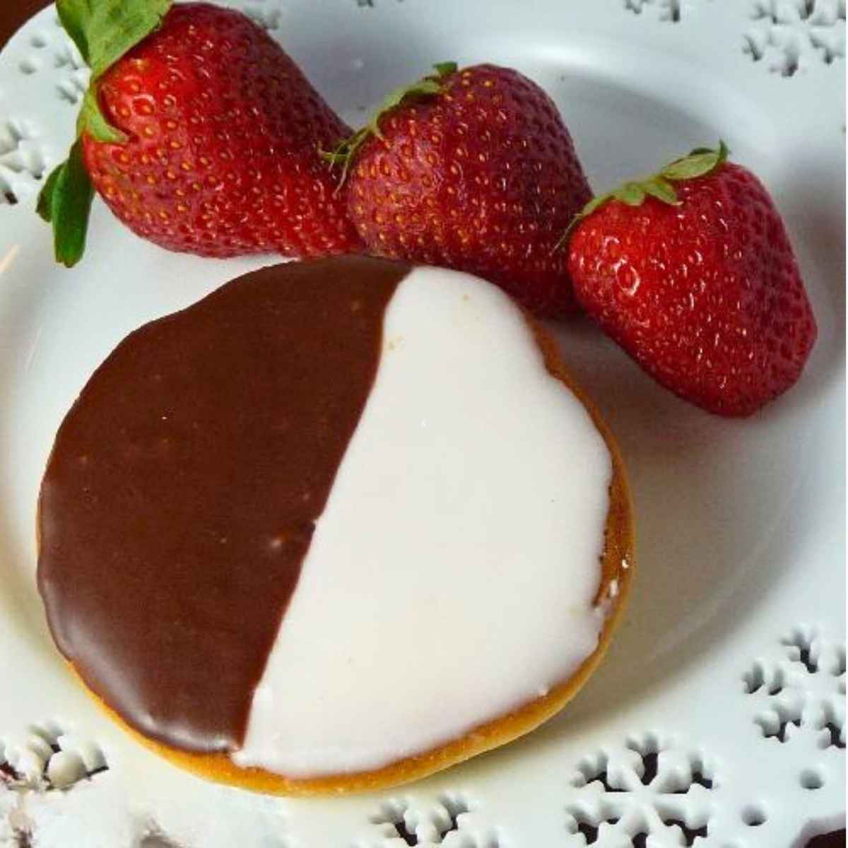 One black and white cookie on a white plate with 3 whole strawberries next to the cookie.